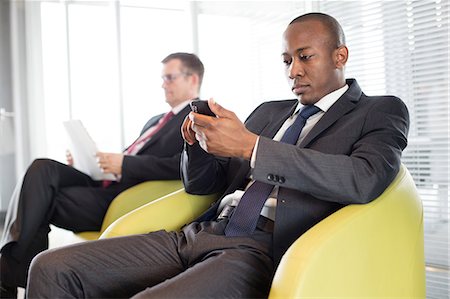 simsearch:693-08769289,k - Young businessman using mobile phone on chair with male colleague in background at office Stock Photo - Premium Royalty-Free, Code: 693-08769344