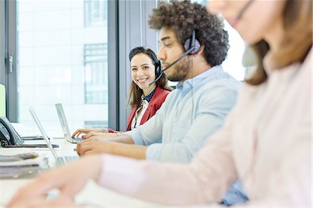 simsearch:693-08769320,k - Portrait of smiling young businesswoman using headset and laptop with colleagues in foreground at office Stock Photo - Premium Royalty-Free, Code: 693-08769598