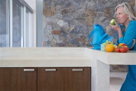 Mature woman leaning on countertop, eating apple, side view Stock Photo - Premium Royalty-Free, Code: 693-03304389