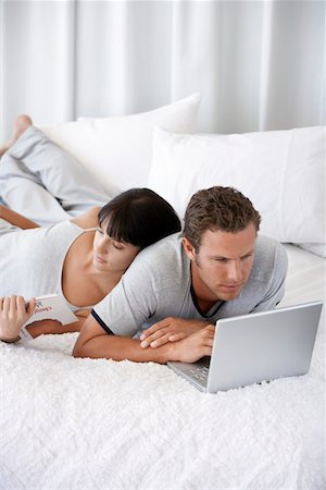 Young couple lying on bed, man using laptop, woman reading Stock Photo - Premium Royalty-Free, Code: 693-03304611