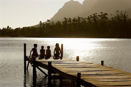 silhouette people sitting on a dock - Three children (7-9) sitting on edge of dock, back view. Foto de stock - Sin royalties Premium, Código: 693-03304765