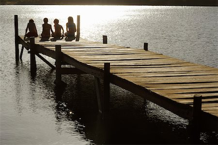 silhouette people sitting on a dock - Four children (7-9) sitting on dock by lake, back view. Foto de stock - Sin royalties Premium, Código: 693-03304766