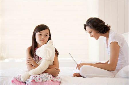 Mother using laptop, daughter cuddling teddy, sitting on bed Stock Photo - Premium Royalty-Free, Code: 693-03306044