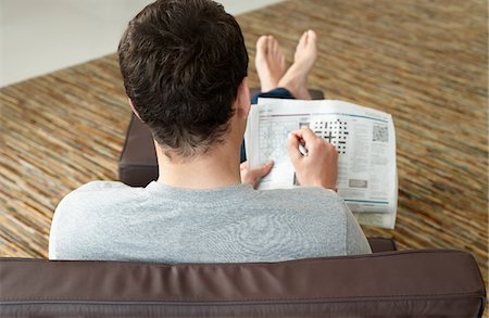 Young man sitting on sofa doing crossword puzzle in newspaper, back view Stock Photo - Premium Royalty-Free, Code: 693-03307432