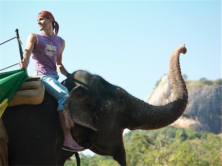 Young man riding elephant, side view, looking over shoulder, mountains in background Stock Photo - Premium Royalty-Free, Code: 693-03307505