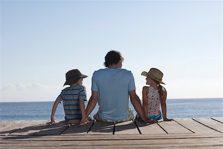 simsearch:693-03309308,k - Father and two children (7-9) sitting on jetty, back view Stock Photo - Premium Royalty-Free, Code: 693-03309306