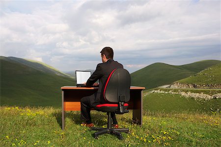 Businessman Sitting at Desk using laptop in mountain field, back view Stock Photo - Premium Royalty-Free, Code: 693-03301666