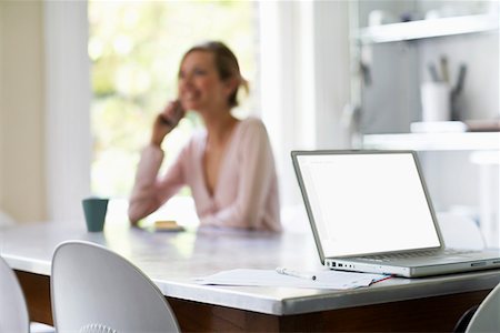 Woman on phone sitting at kitchen table by laptop, focus on laptop Stock Photo - Premium Royalty-Free, Code: 693-03301960