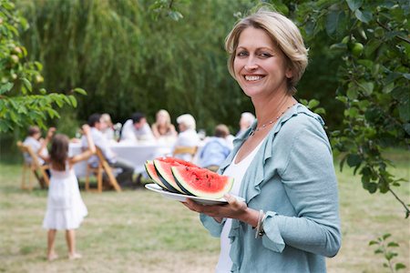 Woman with watermelon slices in garden, family members in background Stock Photo - Premium Royalty-Free, Code: 693-03302741