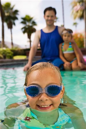 Girl (7-9) in swimming pool, father and sister (7-9) sitting on pool edge in background, front view portrait. Stock Photo - Premium Royalty-Free, Code: 693-03706942