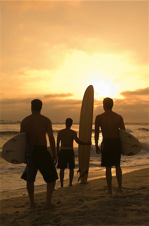 Three surfers standing on beach, holding surfboards, watching sunset, back view Stock Photo - Premium Royalty-Free, Code: 693-03707032