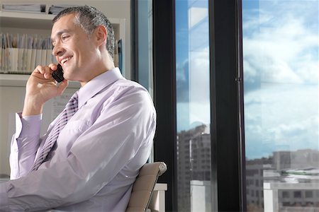simsearch:693-03307925,k - Businessman using mobile phone, sitting in front of window overlooking cityscape Fotografie stock - Premium Royalty-Free, Codice: 693-03707529