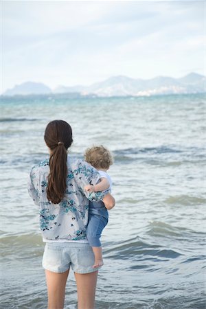 ponytail babies - A lady holding a toddler standing in front of the sea with mountains in the far background Stock Photo - Premium Royalty-Free, Code: 693-03782675