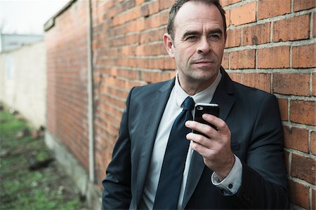 Portrait of businessman standing next to brick wall outdoors, holding cell phone, Germany Fotografie stock - Rights-Managed, Codice: 700-07529283