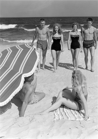 ANNÉES 50 COUPLES EN MAILLOTS DE BAIN MARCHANT VERS LE COUPLE ASSIS SUR SABLE SOUS ÉGIDE Photographie de stock - Rights-Managed, Code: 846-05645934