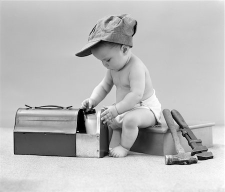 retro baby hat - 1940s BABY IN RAILROAD ENGINEER HAT PULLING MILK BOTTLE FROM LUNCH PAIL WITH HAMMER & WRENCH TOOLS AT SIDE STUDIO Stock Photo - Rights-Managed, Code: 846-05646145