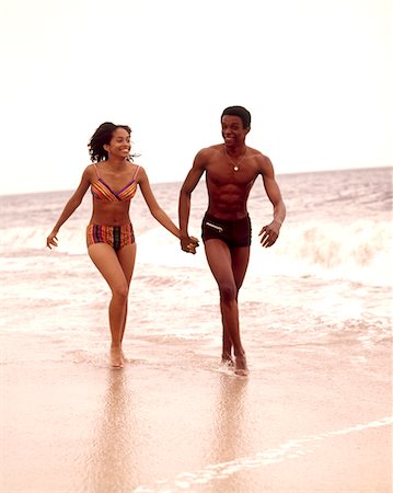 1970s AFRICAN AMERICAN  YOUNG COUPLE WEARING BATHING SUITS RUNNING IN OCEAN SURF Foto de stock - Con derechos protegidos, Código: 846-05646717