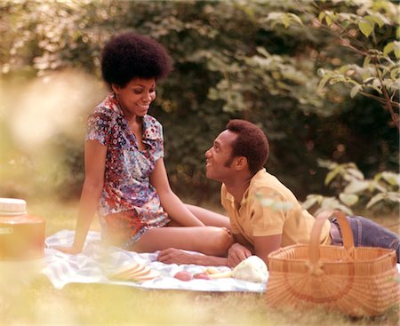 pique-niquer - COUPLE AFRO-AMÉRICAIN ROMANTIQUE DES ANNÉES 1970 L'HOMME PANIER DE PIQUE-NIQUE DE FEMME ASSISE HERBE Photographie de stock - Rights-Managed, Code: 846-05646774