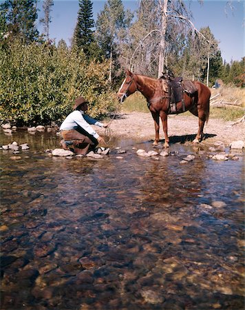 1970s COWBOY IN STREAM KNEELING TRY TO LEAD HORSE INTO WATER Stock Photo - Rights-Managed, Code: 846-05647018