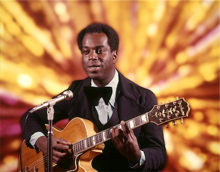 simsearch:846-05647244,k - ANNÉES 1970 AFRO-AMÉRICAIN HOMME ARTISTE CHANTEUSE MUSICIENNE PORTANT TUXEDO PLAYING GUITAR CHANTE EN ARRIÈRE-PLAN LUMIÈRES MICROPHONE OR Photographie de stock - Rights-Managed, Code: 846-05647237