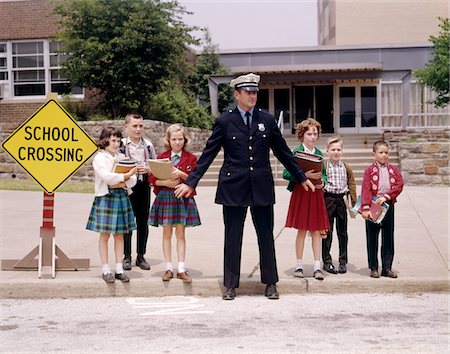 simsearch:846-03164216,k - 1960s MAN TRAFFIC POLICE OFFICER HOLDING BACK GROUP OF ELEMENTARY SCHOOL CHILDREN WAITING AT CURB TO CROSS STREET IN FRONT OF SCHOOL Stock Photo - Rights-Managed, Code: 846-05647451