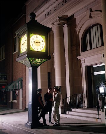 1960s THREE TEENAGE BOYS LOITERING UNDER CLOCK OUTSIDE BANK BUILDING SMOKING CIGARETTES AT NIGHT Stock Photo - Rights-Managed, Code: 846-05647568