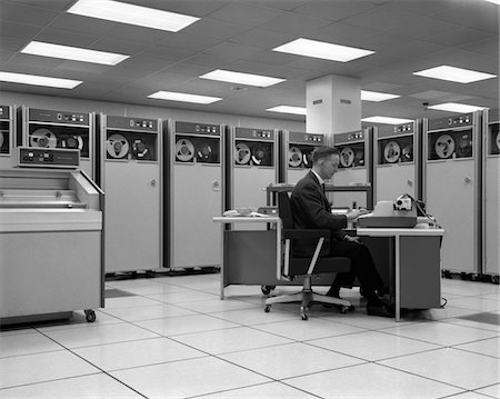 simsearch:846-09012972,k - 1960s MAN TECHNICIAN PROGRAMMING MAINFRAME COMPUTER AT CONTROL ROOM CONSOLE SURROUNDED BY MANY TAPE DRIVE DATA STORAGE MACHINES Stock Photo - Rights-Managed, Code: 846-05647748