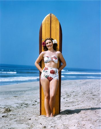 1940s - 1950s SMILING WOMAN WEARING FLORAL PRINT BIKINI BATHING SUIT AND ROSES IN HAIR LEANING AGAINST SURFBOARD ON BEACH Foto de stock - Con derechos protegidos, Código: 846-05647810