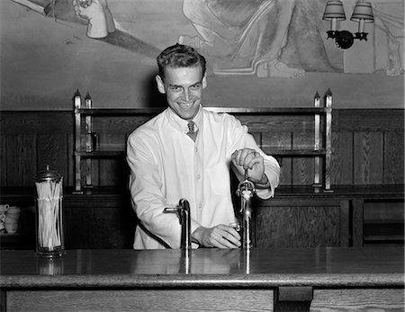 simsearch:846-08512687,k - 1940s SMILING SODA JERK DRESSED IN SHIRT TIE & WHITE SMOCK STANDING BEHIND FOUNTAIN COUNTER FILLING GLASS LOOKING AT THE CAMERA Foto de stock - Con derechos protegidos, Código: 846-06111813