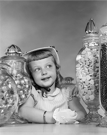 1950s SMILING GIRL WITH BONNET AND WHITE GLOVES SANDWICHED BETWEEN LARGE CANDY JARS ON COUNTER Stock Photo - Rights-Managed, Code: 846-06111939