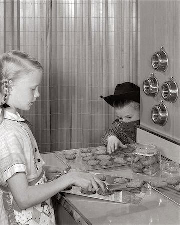 1960s BLOND GIRL TAKING COOKIES OFF TRAY AT KITCHEN COUNTER YOUNGER BOY WITH COWBOY HAT BANDANA MASK REACHES TO STEAL THEM Stock Photo - Rights-Managed, Code: 846-06112000