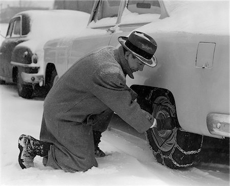 simsearch:846-05648010,k - ANNÉES 1950 HOMME EN MANTEAU & HAT À GENOUX DANS LA NEIGE POUR RÉGLER LES CHAÎNES SUR PNEUS Photographie de stock - Rights-Managed, Code: 846-06112174