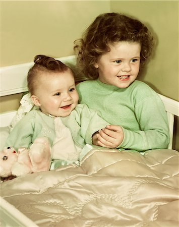1940s 1950s SMILING LITTLE GIRL AND HAPPY BABY SISTER SITTING TOGETHER IN CORNER OF CRIB BED Stockbilder - Lizenzpflichtiges, Bildnummer: 846-08030418