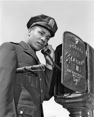 1960s AFRICAN AMERICAN POLICE OFFICER MAKING CALL AT POLICE BOX TELEPHONE NIGHT STICK TUCKED UNDER HIS ARM Stock Photo - Rights-Managed, Code: 846-08140103