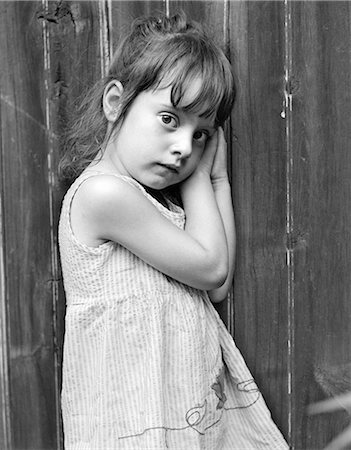 1970s FRIGHTENED SHY UNCERTAIN SAD LITTLE GIRL LEANING AGAINST WOODEN FENCE LOOKING AT CAMERA Foto de stock - Con derechos protegidos, Código: 846-08140113