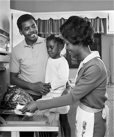 1960s AFRICAN AMERICAN FAMILY IN KITCHEN FATHER AND DAUGHTER WATCHING MOTHER REMOVE ROAST TURKEY FROM OVEN Photographie de stock - Rights-Managed, Code: 846-08226105