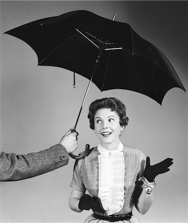 1950s UNSEEN MAN HOLD OUT UMBRELLA TO SMILING WOMAN IN SHIRTWAIST DRESS WHITE RUFFLED BIB FRONT BLACK GLOVES AND CHARM BRACELET Foto de stock - Con derechos protegidos, Código: 846-08226176