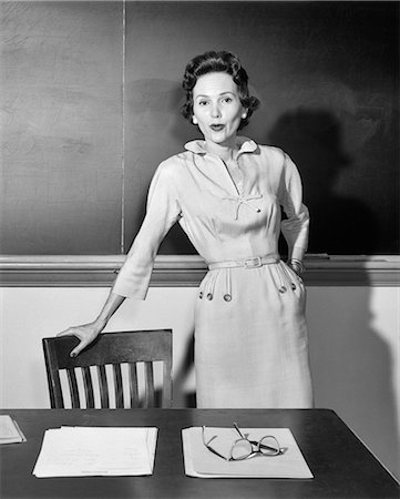 simsearch:846-09161590,k - 1950s WOMAN SCHOOL TEACHER LOOKING AT CAMERA STANDING IN FRONT OF CHALKBOARD IN CLASSROOM LEANING ON CHAIR BY DESK Stock Photo - Rights-Managed, Code: 846-08639582