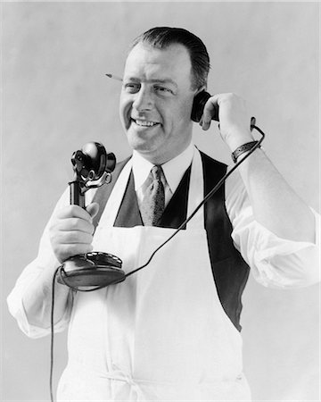1920s MAN IN APRON BUTCHER COOK TALKING ON CANDLESTICK TELEPHONE PENCIL IN EAR Foto de stock - Con derechos protegidos, Código: 846-08721127