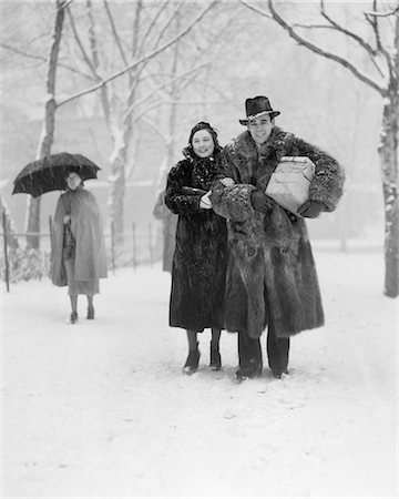 1930s SMILING CHRISTMAS SHOPPING COUPLE WEARING FUR COATS WALKING THROUGH SNOWY CITY PARK CARRYING PACKAGES LOOKING AT CAMERA Foto de stock - Direito Controlado, Número: 846-08721136