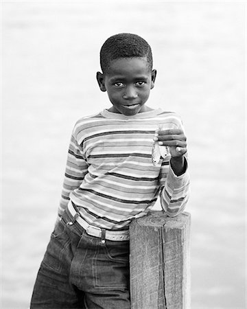 1970s BAHAMIAN BOY IN STRIPED SHIRT WITH SAD FACIAL EXPRESSION LOOKING AT CAMERA ON DOCK HOLDING UP TWO VERY SMALL FISH Photographie de stock - Rights-Managed, Code: 846-09012680