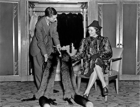 1930s WOMAN IN FUR SALON WEARING FUR COAT SITTING DOWN WHILE SALESMAN SHOWS HER FOX FUR STOLES Photographie de stock - Rights-Managed, Code: 846-09012741