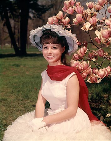 1960s YOUNG WOMAN SITTING BY BLOSSOMING MAGNOLIA TREE WEARING SPRINGTIME HAT RED SCARF WHITE GLOVES AND DRESS LOOKING AT CAMERA Stock Photo - Rights-Managed, Code: 846-09012843
