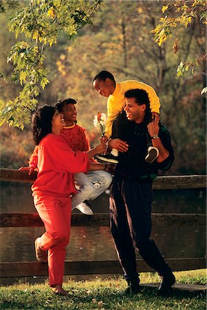 1990s AFRICAN AMERICAN FAMILY STANDING BY FENCE IN AUTUMN BOY HANDING FLOWERS TO MOTHER Stock Photo - Rights-Managed, Code: 846-09012932