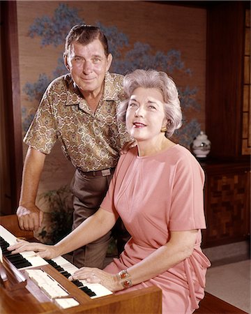 1960S SMILING SENIOR COUPLE TOGETHER AT ELECTRIC HOME ORGAN HUSBAND LOOKING AT CAMERA STANDING BEHIND WIFE WHO IS PLAYING Stock Photo - Rights-Managed, Code: 846-09012953