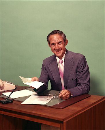 1970s MIDDLE-AGED MAN SITTING AT DESK HOLDING CHECK SMILING LOOKING AT CAMERA Foto de stock - Con derechos protegidos, Código: 846-09012973