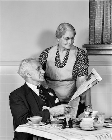 1940s ELDERLY COUPLE MAN WOMAN IN KITCHEN MAN SITTING AT TABLE WOMAN LOOKING OVER HIS SHOULDER READING NEWSPAPER Foto de stock - Con derechos protegidos, Código: 846-09013096