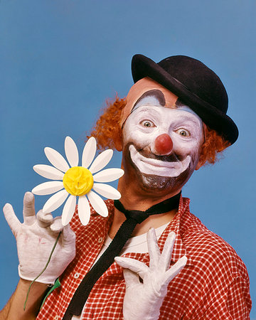 1970s SMILING WIDE-EYED WHITE FACE HOBO CLOWN WITH RED NOSE LOOKING AT CAMERA WEARING A DERBY HAT AND HOLDING A PAPER DAISY Stock Photo - Rights-Managed, Code: 846-09181731