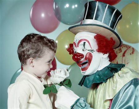 1950s BOY SMILING SMELL SMELLING WHITE ROSE FROM CIRCUS CLOWN TOP HAT RED NOSE BIG SMILE BALLOONS BACKGROUND Stock Photo - Rights-Managed, Code: 846-02794094