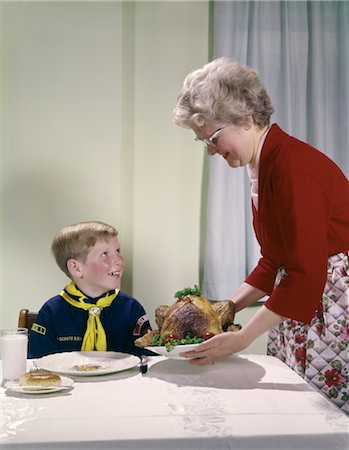 1960s GRANDMOTHER PUTTING TURKEY ON TABLE NEAR BOY IN CUB SCOUT UNIFORM WOMAN GRANDSON FEAST HAPPY OCCASION FOOD Stock Photo - Rights-Managed, Code: 846-02794159